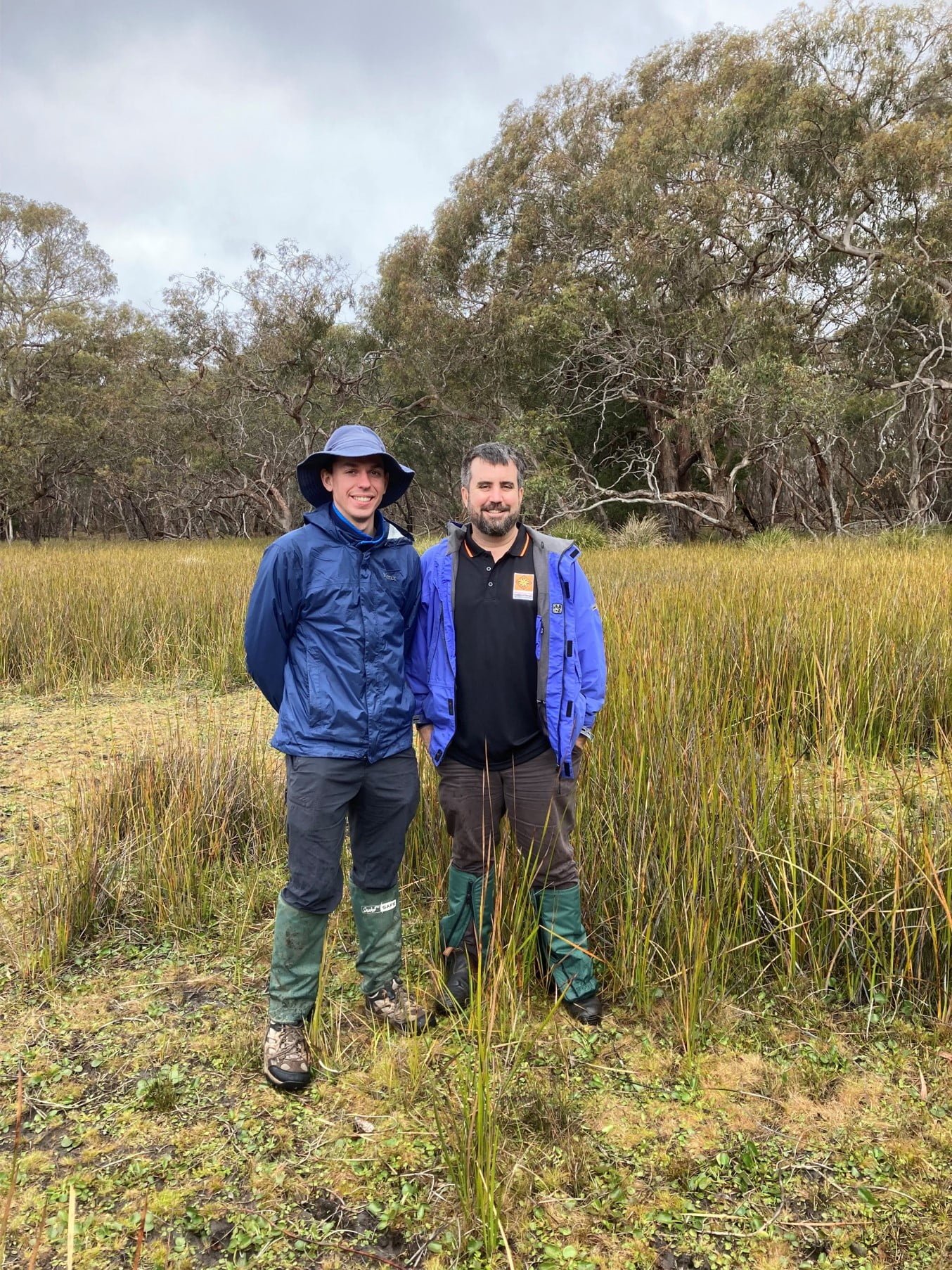 Two men stand side-by-side at a wetland wearing blue rainproof jackets, boots and gators.