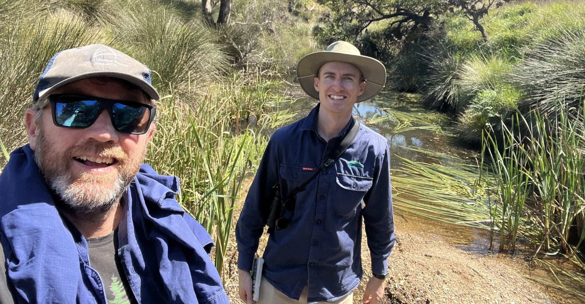Two young men, one with a cap and sunglasses and the other wearing a wide-brimmed hat, smiling at the camera in a selfie photograph taken by a creek in Australia.